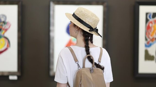 Back view of pretty young Caucasian woman watching and admiring exposition with arts. Exhibition in gallery. Cultural education excursion for students and Museum's Day
