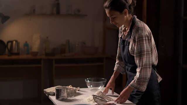 Female baker pouring flour into a metal sifter with hands, carefully preparing baking ingredients on a rustic kitchen table.