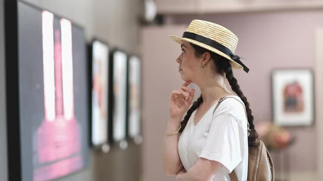 Side view of pretty young Caucasian woman student contemplating exposition with arts. Exhibition in gallery. Cultural education and Museum's Day