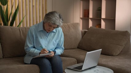 An elderly woman sits on a couch in her living room, taking notes in a notebook while using a laptop. She is focused on her task, demonstrating her engagement with technology