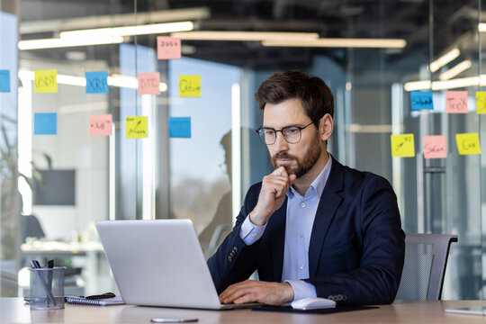 Businessman working on laptop in modern office. Thoughtful professional wearing suit and glasses concentrates on screen. Sticky notes on glass wall suggest brainstorming or project planning