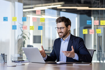 Businessman engaged in virtual meeting using laptop in modern office. Colorful post-its on glass walls indicate brainstorming planning session. Professional setting with confident, happy expression.