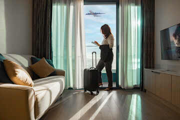Woman with suitcase standing in home or hotel, online check-in in airline's mobile app or on the company's website for self-registration, obtaining boarding pass for quick access to flight at airport.