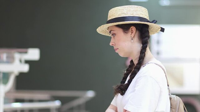 Side view of pretty young Caucasian woman wearing straw hat carefully examines exposition with arts. Exhibition in gallery. Cultural education for students and Museum's Day