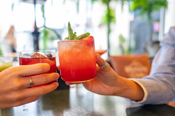 Two people clinking glasses, enjoying red cocktails garnished with mint in a cozy bar setting