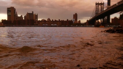 Rainy New York City under Manhattan Bridge in Brooklyn. Pink sunset after rain by river water on waterfront Dumbo promenade, East River. Foggy day weather on riverfront below bridge. Evening twilight.