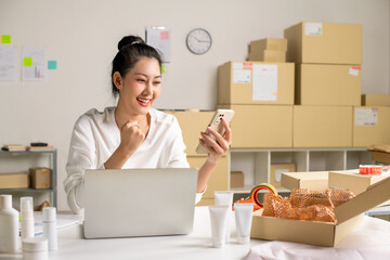Asian e-commerce seller receives order confirmations via smartphone and laptop in a warehouse setting for an online beauty shop.