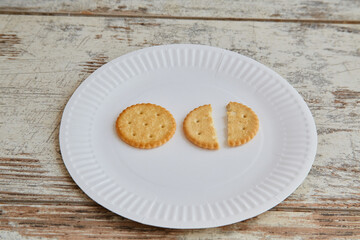 One whole cookie and one cookie broken in half on a white disposable plate on a wooden background close-up, lack of food, rising food prices, meager rations, food shortages