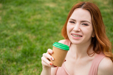 Beautiful young woman with braces on her teeth drinks from a cardboard cup while sitting on the...