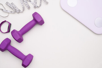 Layout on white surface are purple dumbbells, measuring tape and scales, fitness bracelet, top view. Weight loss and workout concept, fitness accessories