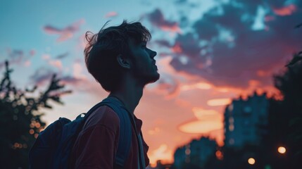 Fototapeta premium A man with a backpack is looking up at the sky. The sky is a beautiful mix of colors, with a purple and orange sunset. The man seems to be lost in thought