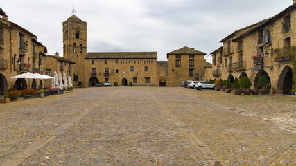 Obraz premium Una panorámica de la Plaza Mayor de Ainsa, Aragón, España, revela una plaza empedrada rodeada de edificios históricos de piedra con arcos en la planta baja. 
