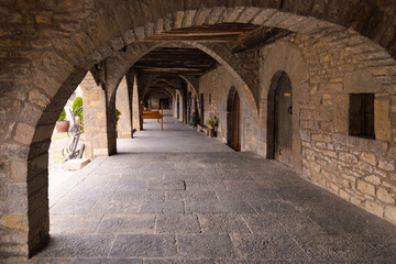 Las arcadas de la Plaza Mayor de Ainsa, Aragón, España, muestran una serie de arcos de piedra que crean un pasillo cubierto y sombreado. 