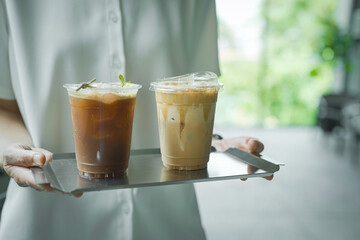 Close up and selective focus hand of employee girl holding tray of ice lychee emericano and cappuccino at cafe.