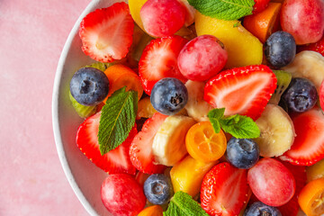 Summer fresh fruit salad in a bowl on pink background. Top view. Close up. Healthy food for breakfast. Mixed strawberries, grapes, banana, kiwi, blueberries, peach, kumquat and mint for diet lunch