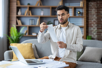 Stressed man looking at bills while working on laptop in living room