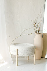 Interior design of living room with white plastered walls, stylish white jute pouf with wooden legs, natural treed twigs in clay vase and big floor mirror. Scandinavian modern design home decor.