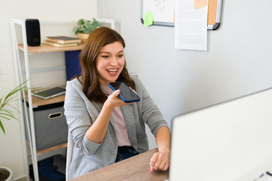 Cheerful young businesswoman using smartphone for a voice command or chat in a bright startup office environment, displaying productivity and technology