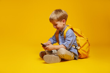 Happy adorable little boy sitting on studio floor with yellow backpack, playing on smartphone and smiling, satisfied with children mobile application. Isolated on orange background.