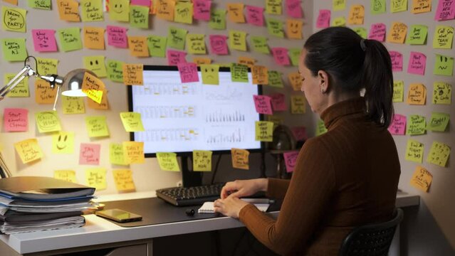 Young woman adds colorful note to her computer screen while working late at night, surrounded by sticky notes covering the walls. Time organization and planning concept