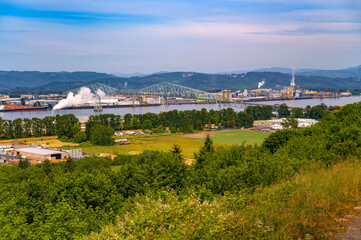 View of the Lewis and Clark Bridge spanning the Columbia River between the states of Washington and Oregon, surrounded by industrial facilities and lush green landscape.