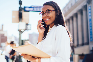 Portrait of cheerful dark skinned female employee holding folder in hands while calling with friends on smartphone device standing in urban setting.Successful lawyer in white shirt talking on cellular