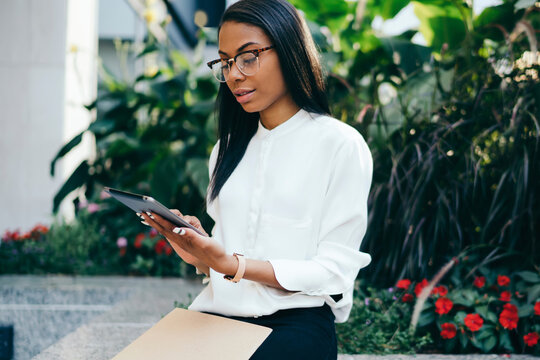 Pensive african american young woman in formal wear reading financial news on modern tablet connected to 4G internet.Dark skinned female office worker checking mail on touch pad sitting outdoors