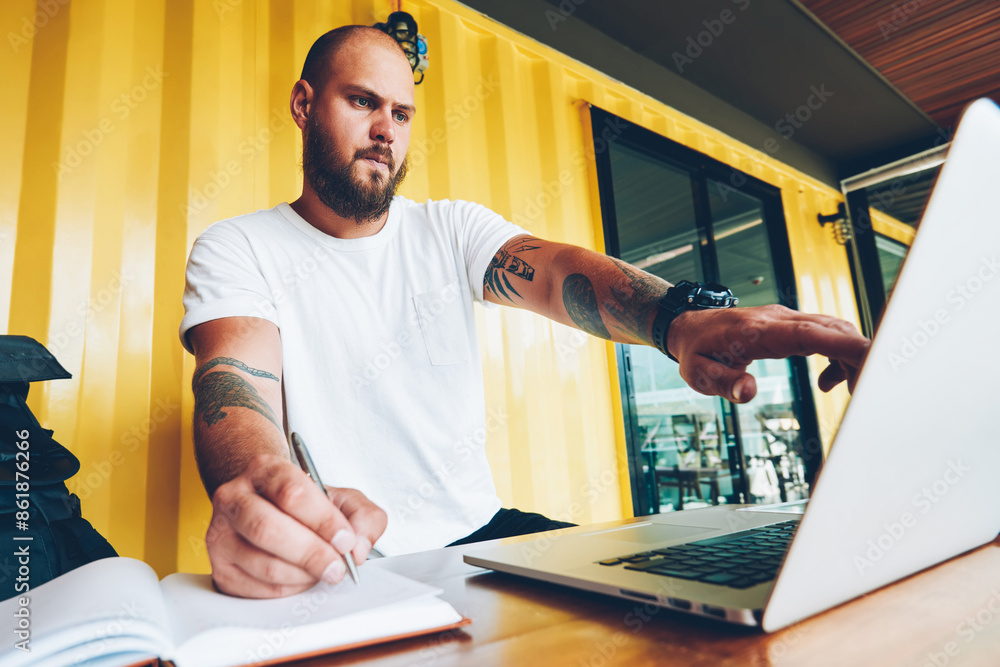 Poster clever caucasian male student copying information from web page to notebook doing homework using lap