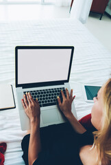 Fototapeta premium Top vie of female freelancer keyboarding on laptop computer working sitting on comfortable bed, hipster girl using netbook with mock up screen typing publication for blog doing distance job at home