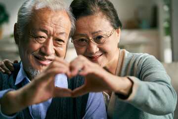 happy loving senior asian couple forming a heart shape with hand while video chatting with friends and family