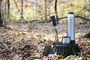 Thermos and hunting knife. The concept of tourism, survival and hunting in the autumn in forest. a thermos of tea, a cup and a knife, on an old tree stump. dry leaves in the park. rest at nature.