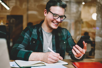 Cheerful hipster guy in spectacles laughing on new message from friend holding smartphone while writing homework, positive prosperous male freelancer working in cafe interior using modern technology