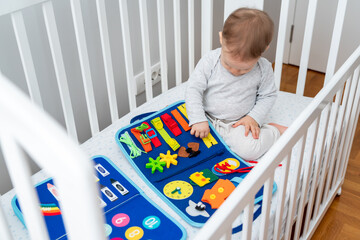 Baby playing with montessori busy book sitting in crib. Concept of smart books and modern toys