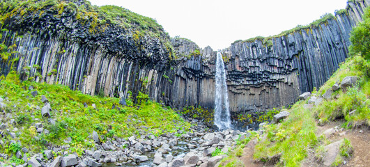 Svartifoss, known for its distinct basalt columns, cascades down the rugged cliff face surrounded...