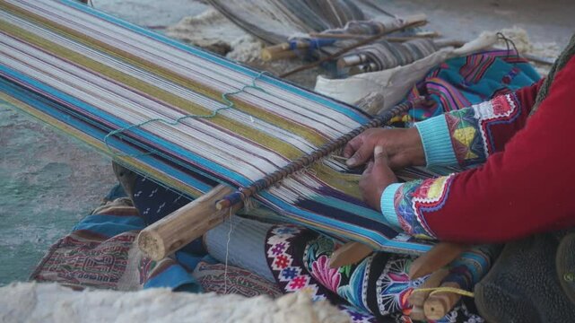 A woman is sitting on the ground and working on a piece of fabric. The fabric is striped and has a blue and white pattern. The woman is using a wooden stick to weave the fabric. The scene has a calm
