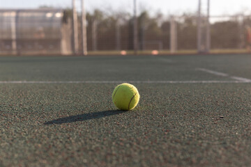 yellow tennis ball sits on a green tennis court, with a blurred background of a tennis net and trees