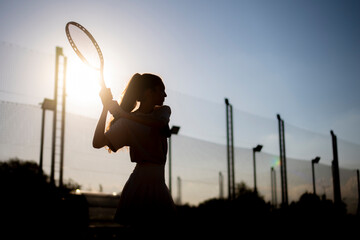 silhouette of a female tennis player executing a backhand swing during a sunset match on an outdoor...