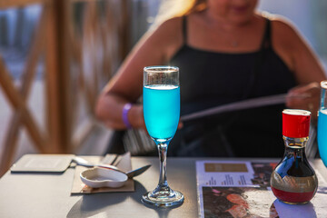 Close-up view of table with blue drink in glass and blurred woman's silhouette in background at outdoor restaurant. Curacao.