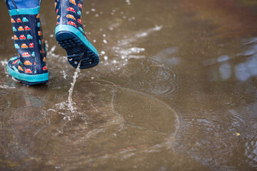 The kid in funny rubber boots walking along a puddle on the street after the rain. A pair of colorful rubber boots in a large pool with drops of water. The boy had fun after rain in the open air