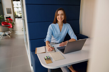 A professional woman is smiling, using a laptop, and taking notes in a modern office environment