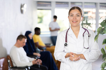 Smiling polite young female doctor in white coat standing indoors against blurred background of busy clinic lobby with healthcare workers and patients..