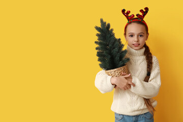 Cute little girl wearing reindeer horns with Christmas tree in pot on yellow background
