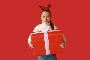 Cute little girl in reindeer horns with Christmas gift on red background