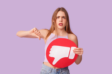 Beautiful young displeased woman with dislike icon showing thumb-down gesture on purple background