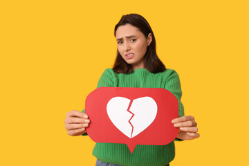 Beautiful young displeased woman with heart dislike icon on yellow background