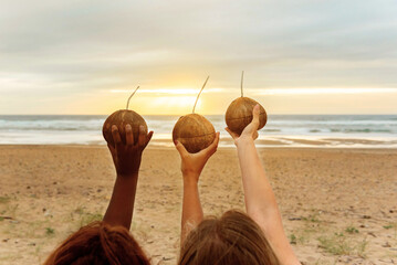 three unrecognizable young women of different races and skin tones holding in their raised hand a drink served in a coconut during a beach party at sunset.