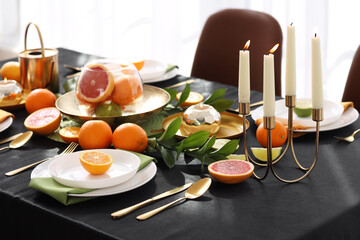 Beautiful table setting with citrus fruits, burning candles and black tablecloth in living room