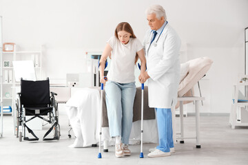 Mature doctor helping female patient with crutches in clinic