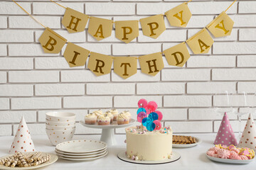 Table with cake, sweets and decor for birthday party against white brick wall
