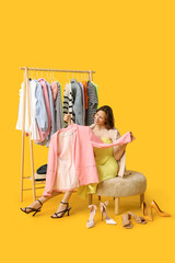 Beautiful young woman near rack with different clothes choosing blazer and shoes on yellow background